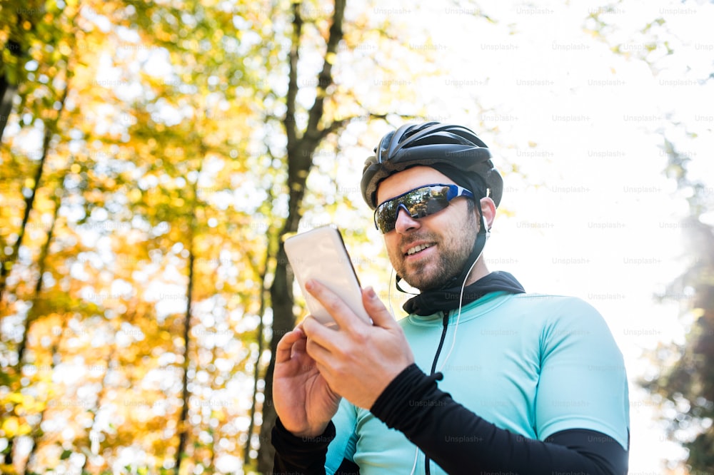 Jovem esportista bonito andando de bicicleta do lado de fora na natureza ensolarada do outono. Segurando o telefone inteligente, ouvindo música.
