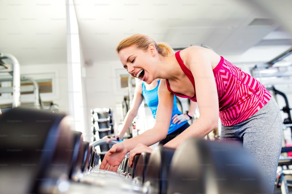 Gros plan de deux femmes attrayantes en forme dans la salle de sport riant, rangée de poids