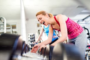 Close up of two attractive fit women in gym laughing, row of weights