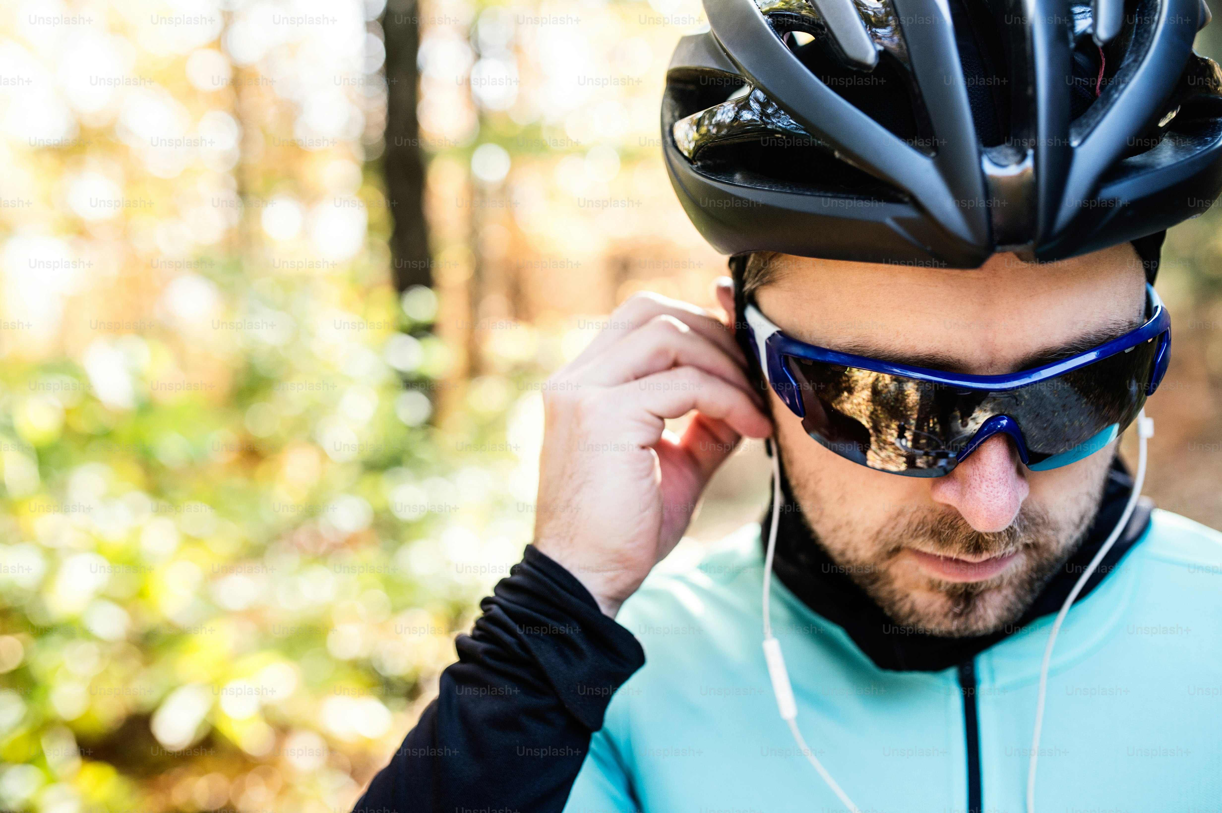 Joven deportista guapo montando su bicicleta al aire libre en la naturaleza soleada del otoño, poniéndose auriculares en los oídos, vista trasera