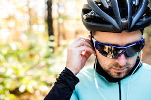 Joven deportista guapo montando su bicicleta al aire libre en la naturaleza soleada del otoño, poniéndose auriculares en los oídos, vista trasera