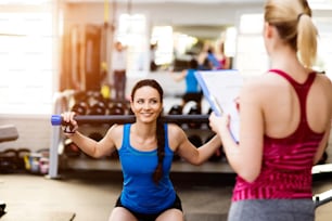 Close up of an attractive fit woman in blue singlet exercising in a gym with her personal trainer writing into exercise plan on clipboard