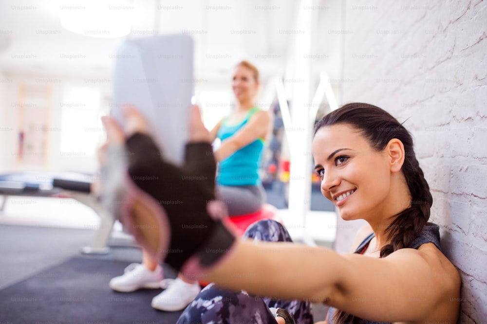 Mujer sonriente y atractiva en forma en el gimnasio sentada en un piso sosteniendo un teléfono inteligente y tomando selfie contra la pared de ladrillo blanco