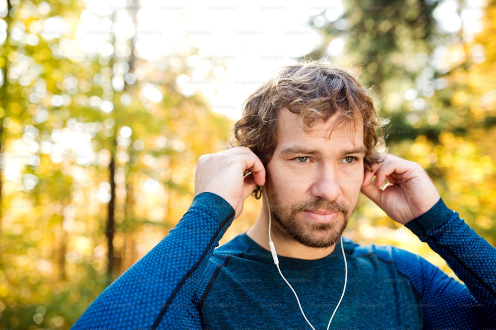 Young handsome runner putting earphones in his ears, listening music, outside in sunny autumn nature
