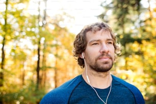 Young handsome runner with earphones in his ears, listening music, outside in sunny autumn nature