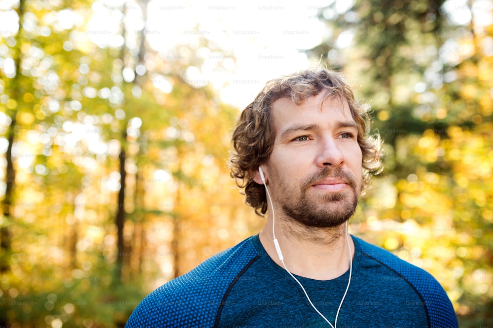 Young handsome runner with earphones in his ears, listening music, outside in sunny autumn nature