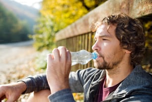 Junger, gutaussehender Läufer mit Wasserflasche auf einer Brücke sitzend, trinkend, ruhend