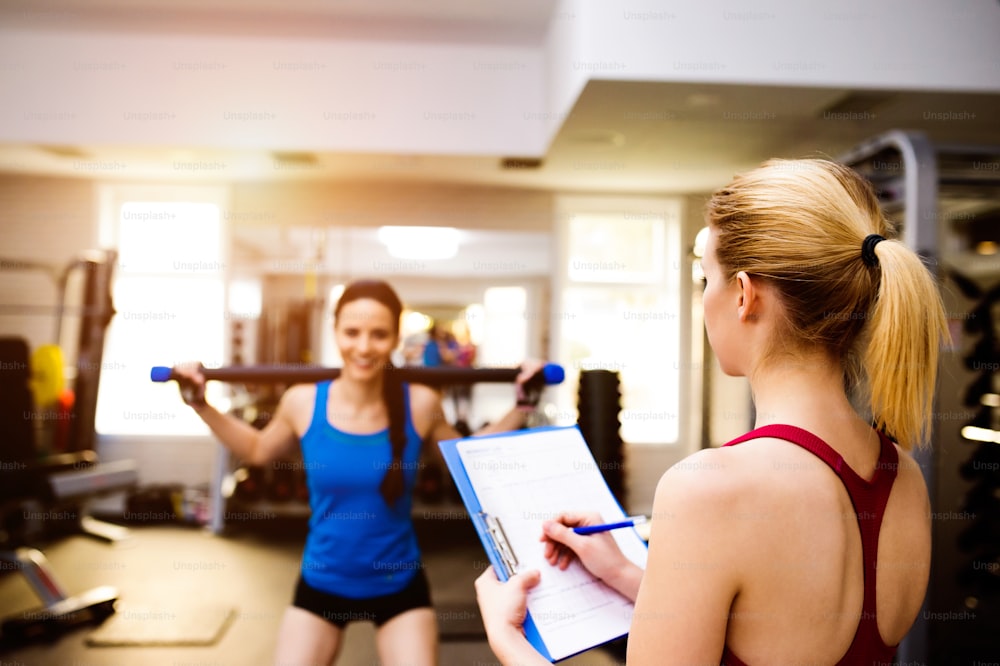 Mulher de singlet azul exercitando agachamentos com barra em uma academia com seu personal trainer escrevendo em plano de exercícios na prancheta