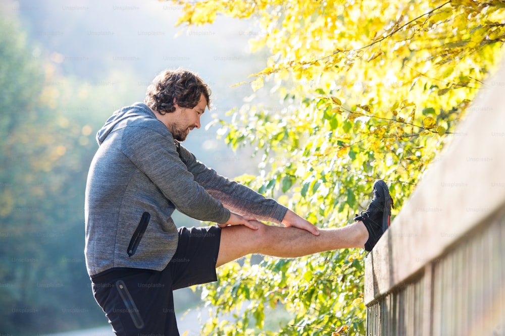 Handsome young runner warming up and stretching his legs on a bridge. Sunny autumn nature.