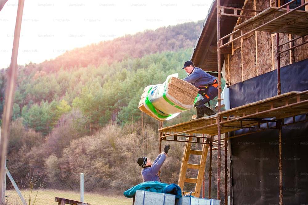 Construction worker standing on scaffold thermally insulating house facade with glass wool. Other worker passing him another bale up on platform.