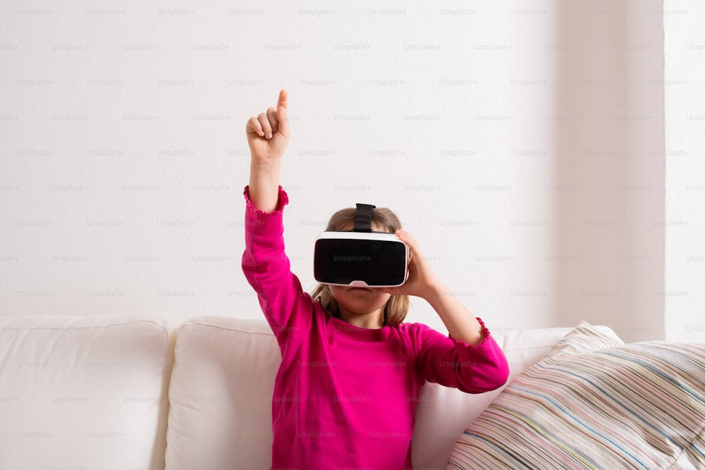 Cute little girl in pink t-shirt wearing virtual reality goggles. Studio shot, white couch, copy space