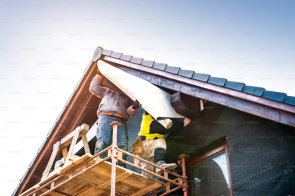 Construction workers standing on scaffold thermally insulating house facade with glass wool and foil.