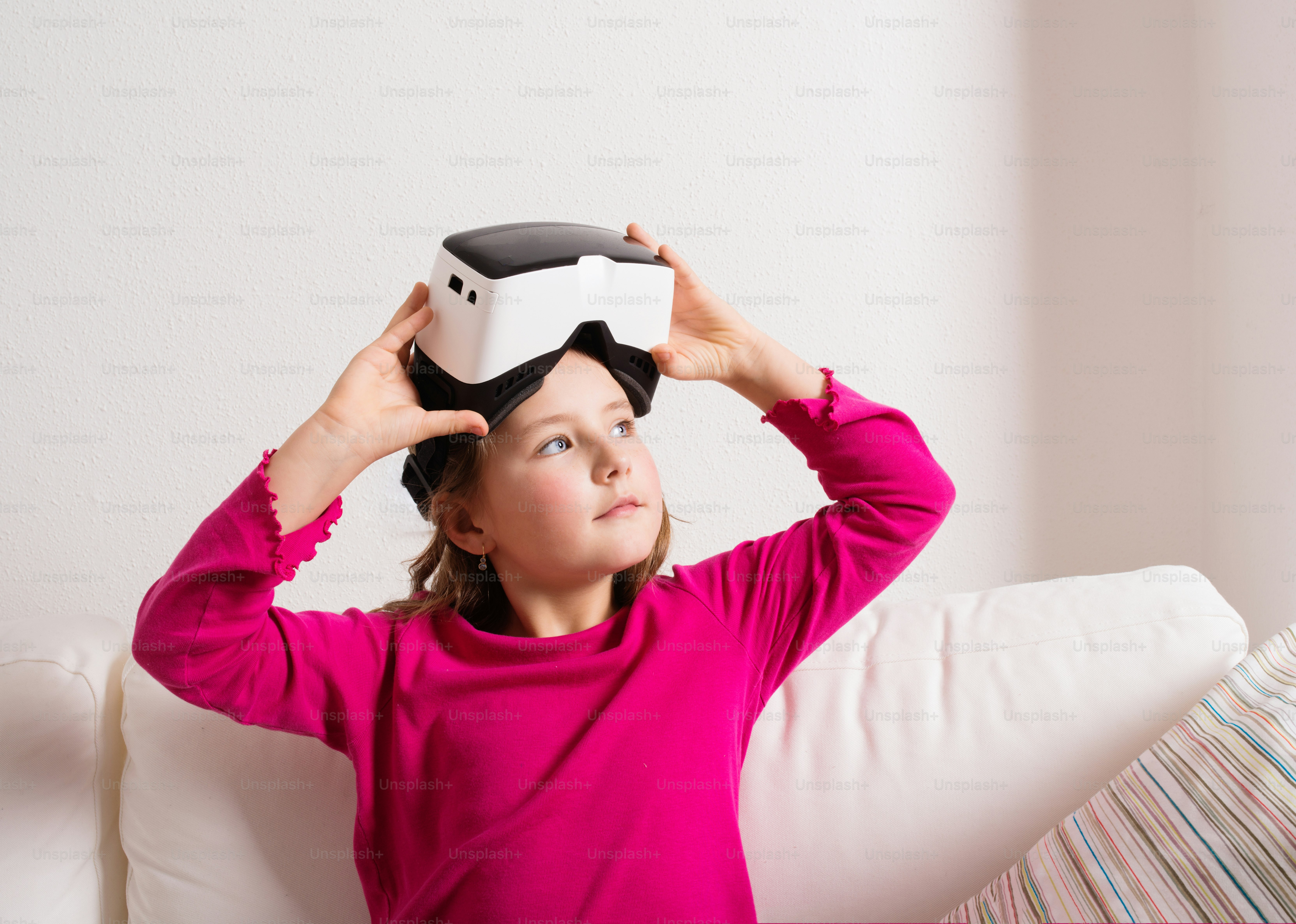Cute little girl in pink t-shirt wearing virtual reality goggles. Studio shot, white couch, copy space