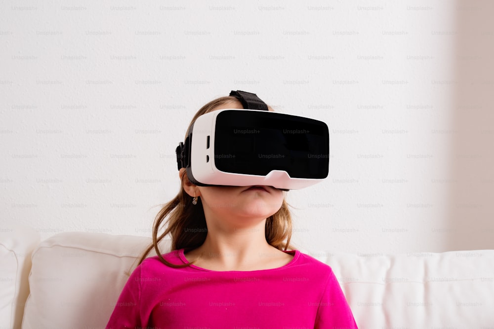 Linda niña con camiseta rosa con gafas de realidad virtual. Foto de estudio, sofá blanco, espacio de copia