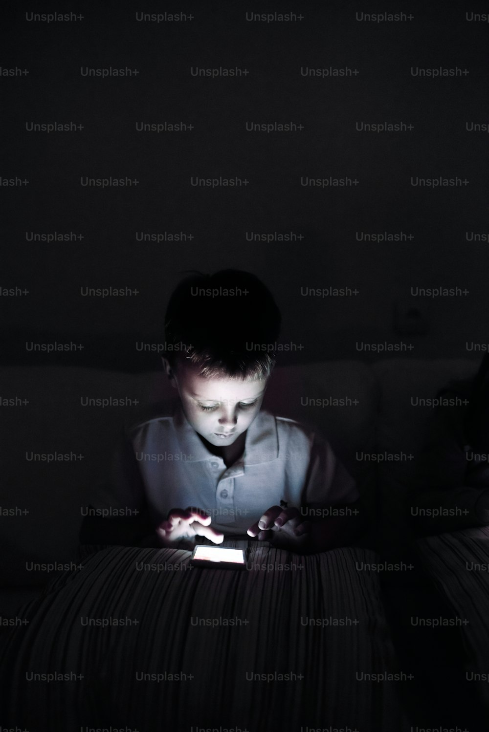 Niño pequeño jugando con un teléfono inteligente, sentado en el sofá en una habitación oscura por la noche.