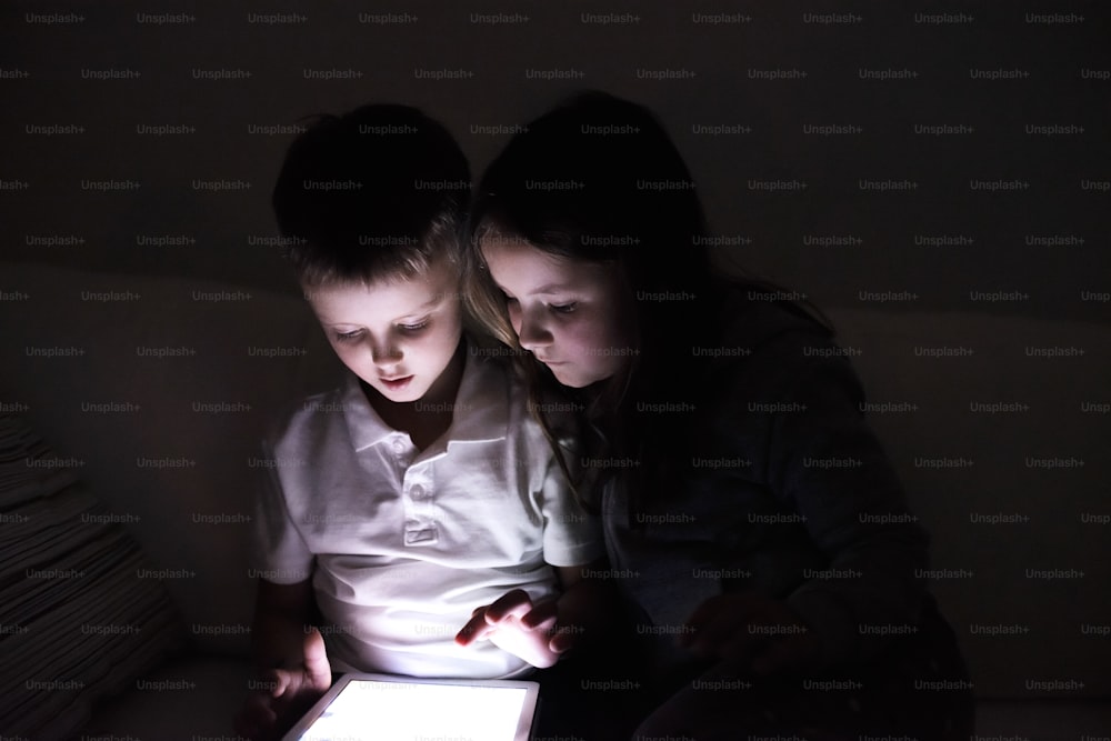 Two children playing with tablet, sitting on sofa in dark room at night.
