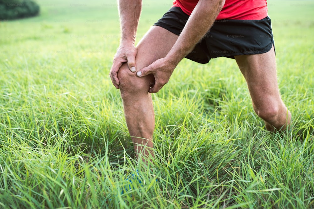 Unrecognizable active senior runner outside in field. Man with injured knee. Close up. Green sunny summer nature.