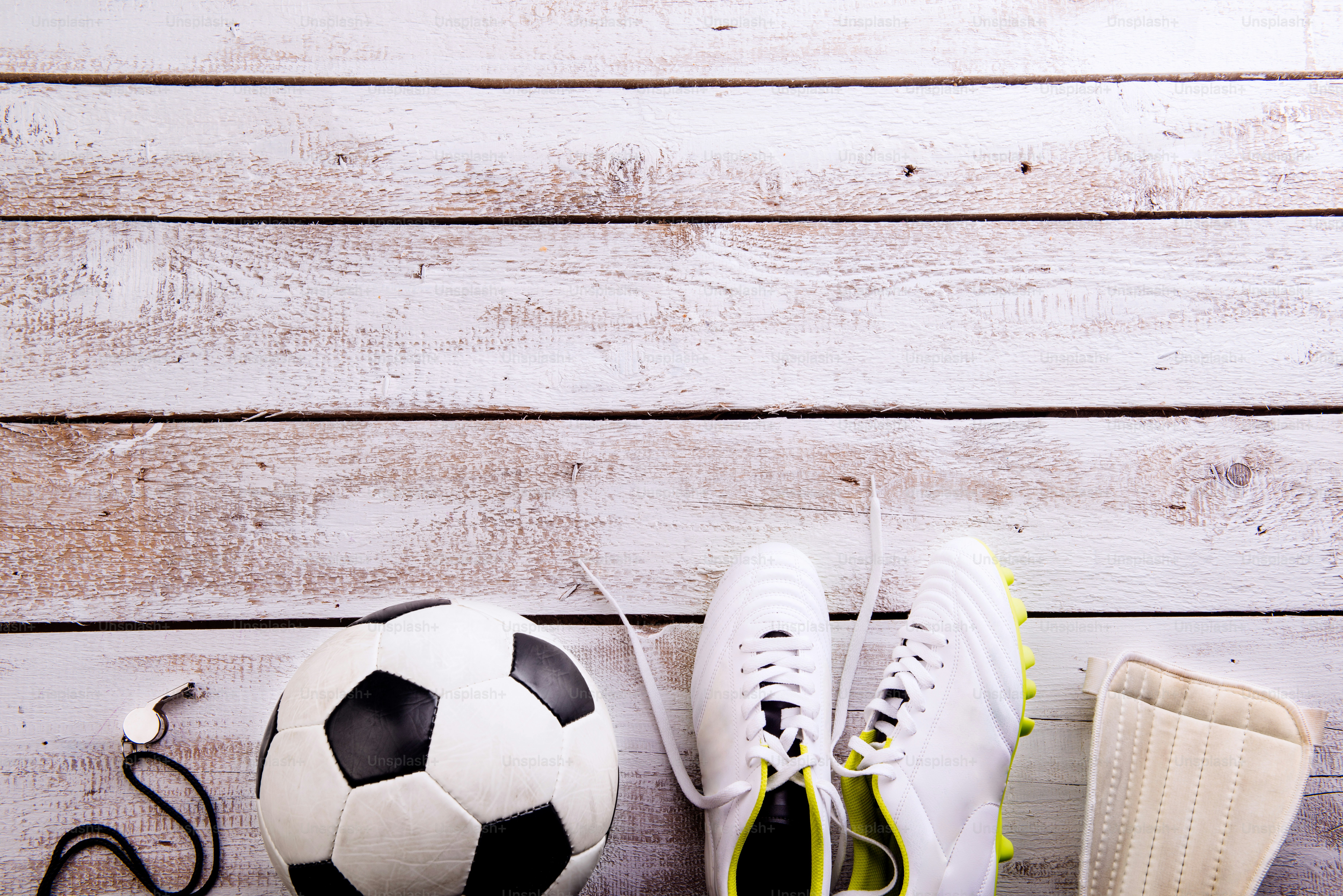 Soccer ball, cleats, protectors and whistle against wooden floor, studio shot on white background. Flat lay, copy space