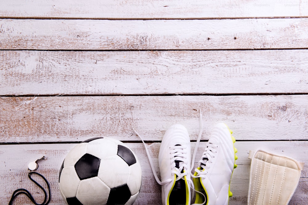 Soccer ball, cleats, protectors and whistle against wooden floor, studio shot on white background. Flat lay, copy space