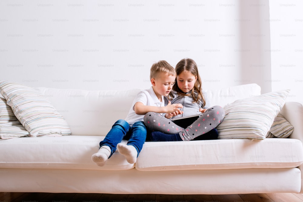 Niña y niño sentados en el sofá con una tableta. Niños felices jugando en el interior.