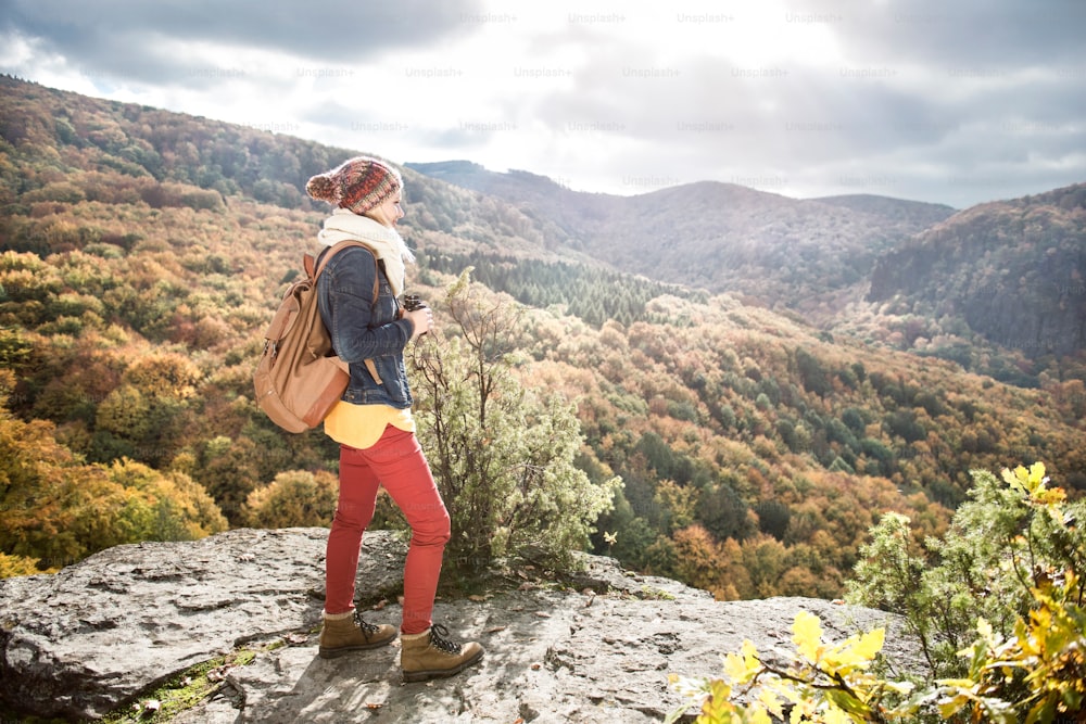 Beautiful woman with backpack, holding binoculars, standing on a rock against colorful autumn forest, rear view