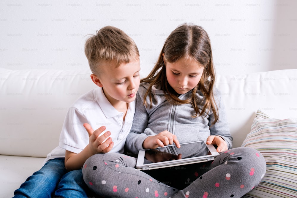 Petite fille et garçon assis sur un canapé avec une tablette. Des enfants heureux qui jouent à l’intérieur.