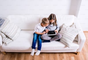 Little girl and boy sitting on sofa with a tablet. Happy children playing indoors.