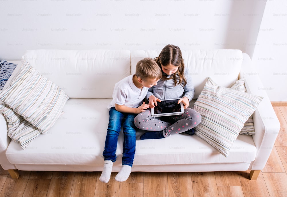 Menina e menino sentados no sofá com um tablet. Crianças felizes brincando dentro de casa.