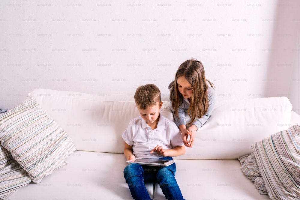 Niña y niño sentados en el sofá con una tableta. Niños felices jugando en el interior.