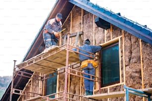 Des ouvriers de la construction debout sur un échafaudage isolant thermiquement la façade de la maison avec de la laine de verre.