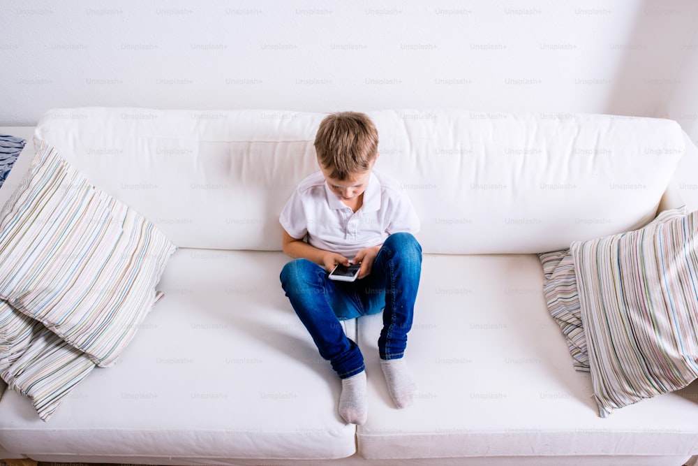 Little boy sitting on sofa with a smart phone. Happy child playing indoors.