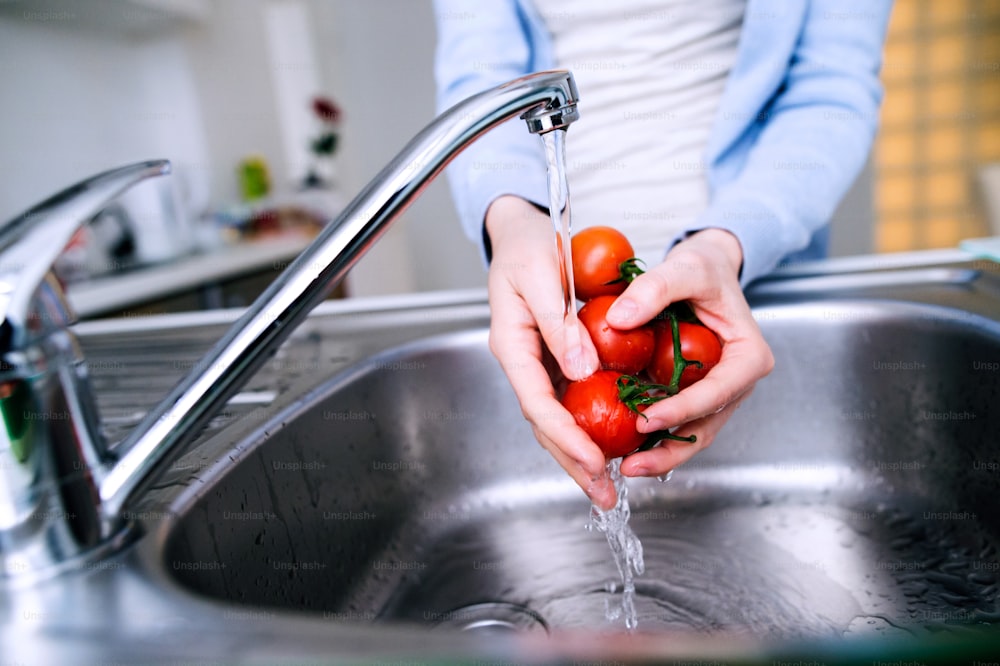 チェックの青いシャツを着た見分けのつかない年配の女性の手が、蛇口の下でトマトを洗っている。朝食の準備。