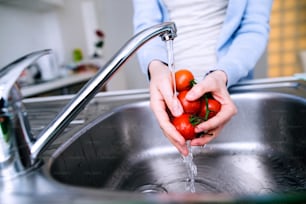 Hands of unrecognizable senior woman in checked blue shirt washing tomatoes under te tap. Preparing breakfast.
