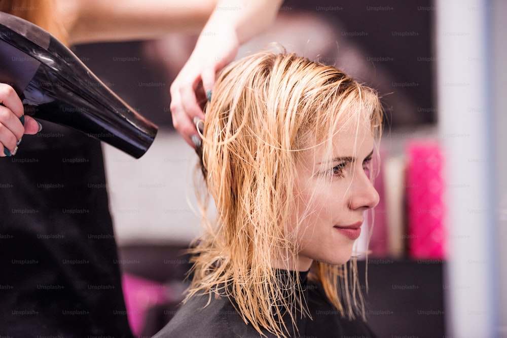 Hands of unrecognizable professional hairdresser drying hair of her client, new haircut, blonde female customer.
