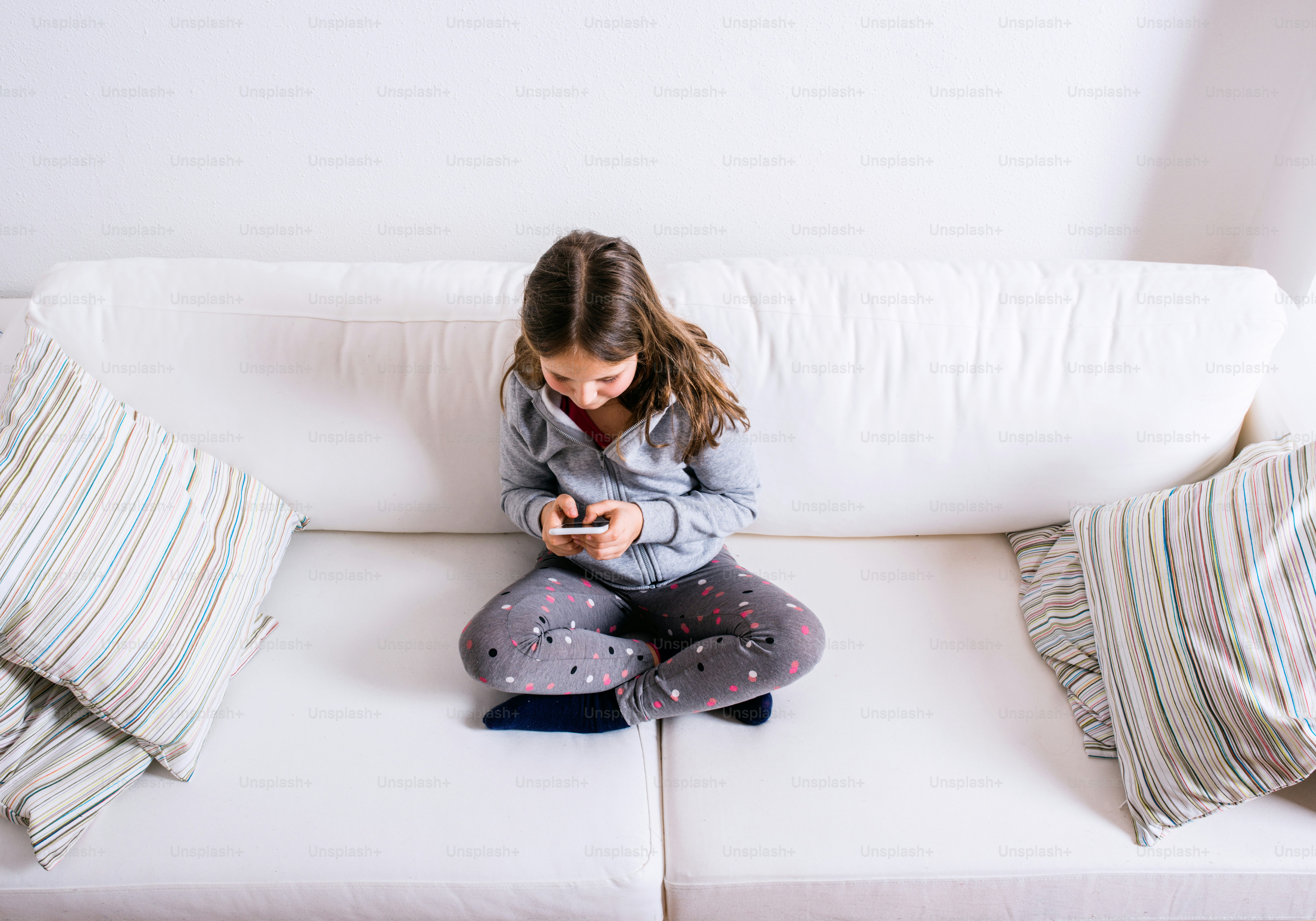 Little girl sitting on sofa with a smart phone. Happy child playing indoors.