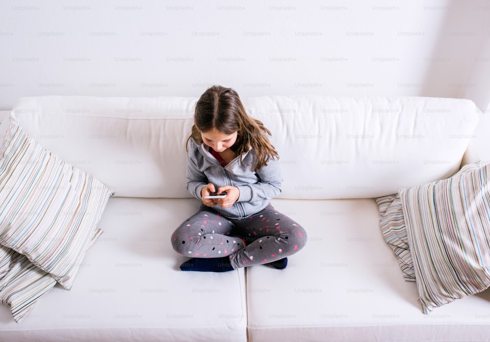 Bambina seduta sul divano con uno smartphone. Bambino felice che gioca al chiuso.