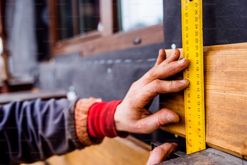 Hands of unrecognizable construction worker thermally insulating house, doing wooden facade, measuring board with yellow tape measure