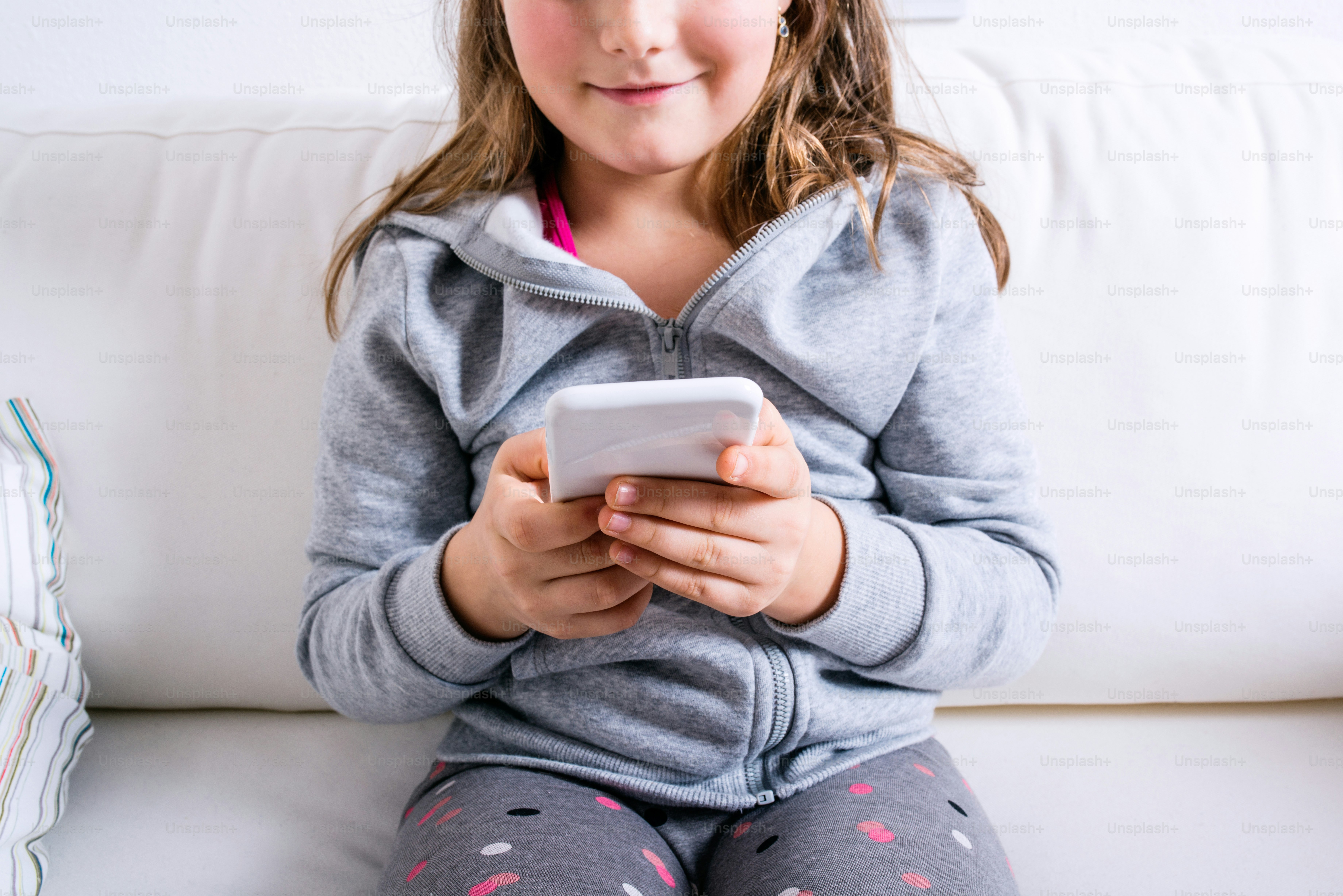 Unrecognizable little girl sitting on sofa with a smart phone. Happy child playing indoors.