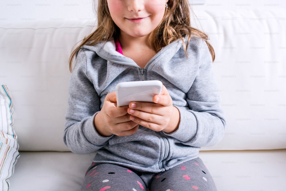 Petite fille méconnaissable assise sur un canapé avec un téléphone intelligent. Enfant heureux jouant à l’intérieur.