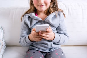 Petite fille méconnaissable assise sur un canapé avec un téléphone intelligent. Enfant heureux jouant à l’intérieur.