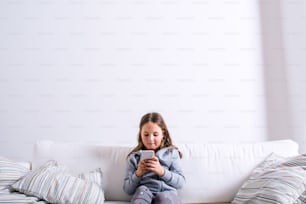 Menina sentada no sofá com um telefone inteligente. Criança feliz brincando dentro de casa.