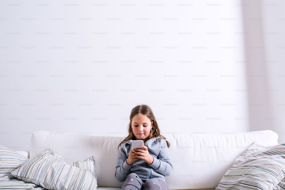 Kleines Mädchen sitzt mit einem Smartphone auf dem Sofa. Glückliches Kind, das drinnen spielt.