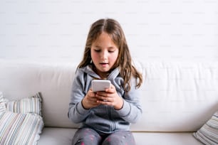 Niña sentada en el sofá con un teléfono inteligente. Niño feliz jugando en el interior.