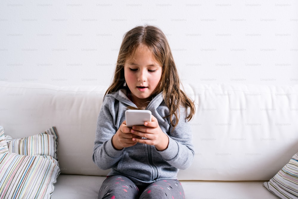 Menina sentada no sofá com um telefone inteligente. Criança feliz brincando dentro de casa.