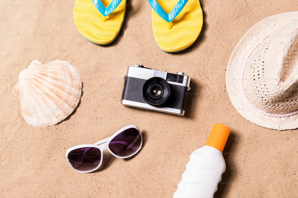 夏休みの構図は、カメラ、黄色いビーチサンダル、帽子、サングラス、日焼け止めクリームなど、ビーチで。砂の背景、スタジオショット、フラットレイ。