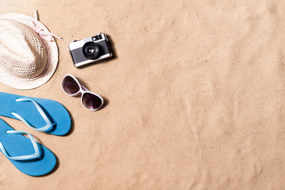 여름 휴가 구성으로 파란색 플립 플롭 샌들, 모자, 선글라스 및 복고풍 스타일의 카메라가 해변에 놓여 있습니다. 모래 배경, 스튜디오 촬영, 플랫 레이. 공간을 복사합니다.