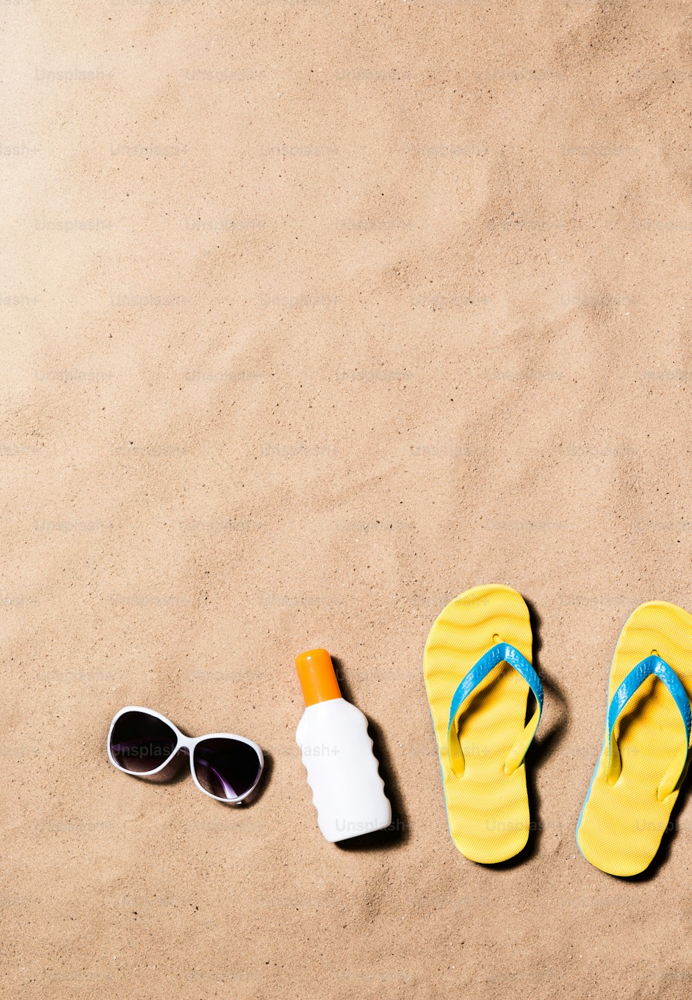 Composizione per le vacanze estive con un paio di sandali infradito gialli, occhiali da sole e crema abbronzante su una spiaggia. Sfondo spiaggia di sabbia, scatto in studio, flat lay. Copia spazio.