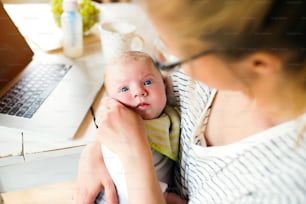 Close up of unrecognizable mother holding her baby son in the arms, working on laptop, milk in the bottle