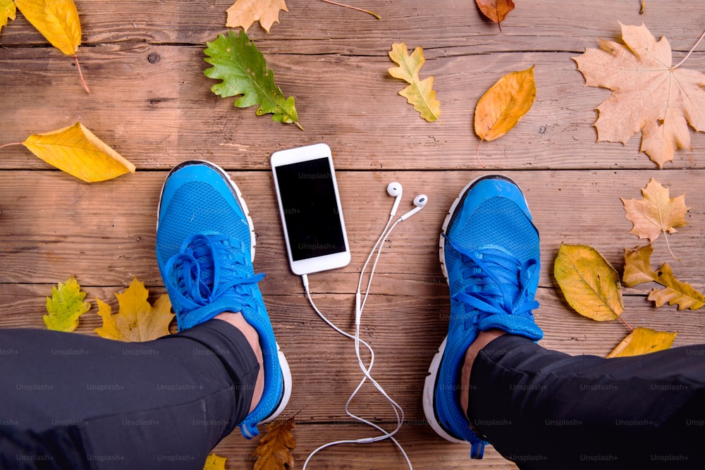 Legs of unrecognizable runner in blue sports shoes. Smart phone and earphones. Colorful autumn leaves. Studio shot on wooden background.