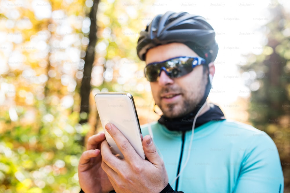 Young handsome sportsman riding his bicycle outside in sunny autumn nature. Holding smart phone, listening music.